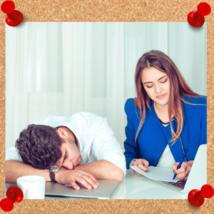 A male and a female employee sit at a desk while the male sleeps and the female works.