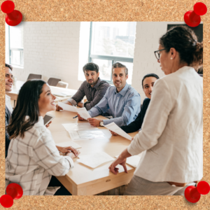 Image depicts a work team gathered around a conference table as one woman stands at the head.