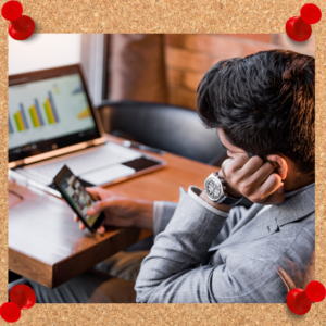 Image depicts man playing on his phone while seated at his desk. His laptop computer is open to a series of bar graphs, indicating that he has yet to stop procrastinating on his work.