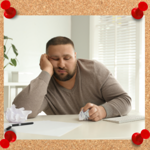 Man appears to be sleeping while he should be working. There is paper and a pen on the table or surface where he rests with a crumpled up page in his fist.