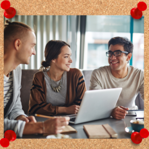 Image depicts a group of three employees chatting around a conference table with a laptop between them.